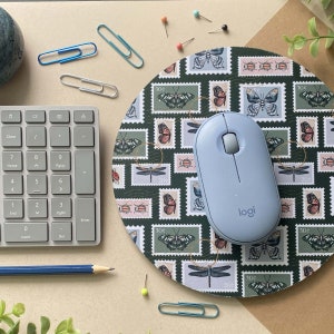 Pode incluir: Um rato de computador sem fio azul com a palavra "logi" impressa nele, sentado em um mousepad redondo verde com um padrão de borboletas e libélulas em selos postais.