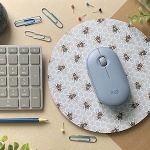Pode incluir: Um tapete de rato redondo com um fundo azul claro e um padrão de favo de mel. O tapete de rato está coberto de pequenas abelhas amarelas. Um rato de computador sem fios azul claro com a palavra "logi" impressa nele está sentado no tapete de rato.
