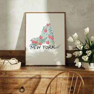 May include: A framed print of the state of New York, outlined in a floral design with red apples and green leaves. The print is on a white background and has the words "NEW YORK" written below the outline.