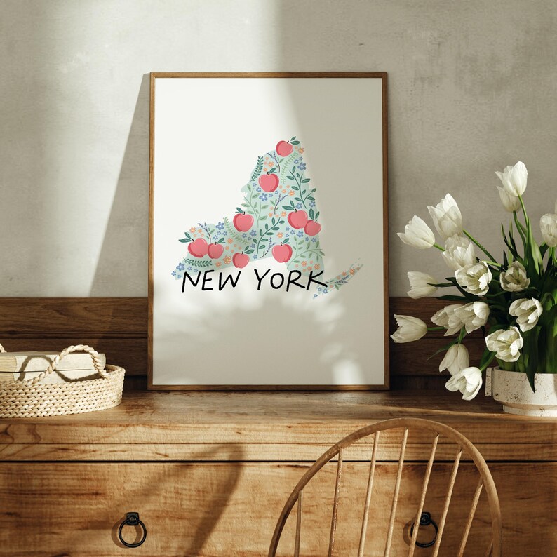 May include: A framed print of the state of New York, outlined in a floral design with red apples and green leaves. The print is on a white background and has the words "NEW YORK" written below the outline.