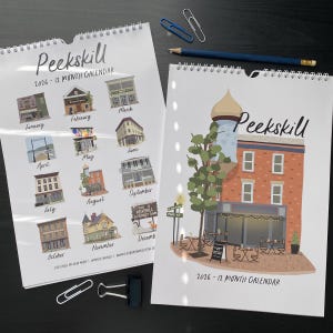 May include: Two spiral-bound calendars with illustrations of buildings. One calendar shows a monthly view with building images. The other features a detailed illustration of a building with the text "Peekskill" and "2026-12 MONTH CALENDAR".
