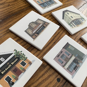 May include: Set of six white ceramic coasters featuring illustrations of different buildings. Each coaster has a unique building design with details like windows, doors, and signage. The coasters are ideal for protecting surfaces from drink spills.