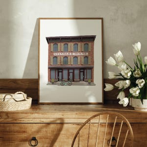 May include: A framed print of a brick building with the text "STANDARD HOUSE." The building has a brown roof, white windows, and a brown door. Two bicycles are parked in front of the building.