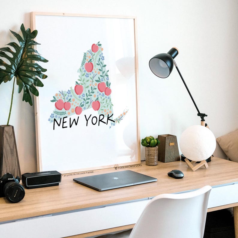 May include: A framed print of the state of New York, outlined in green leaves and filled with red apples and white flowers. The words "NEW YORK" are written in black below the outline.