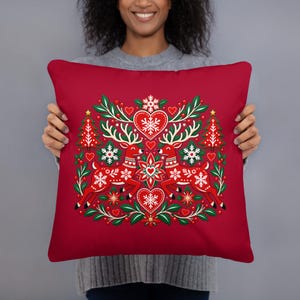 May include: A square red throw pillow featuring a central graphic of two stylized red reindeer facing each other, surrounded by white snowflakes, red hearts, green foliage, and small red berries, with two red Christmas trees on the sides.