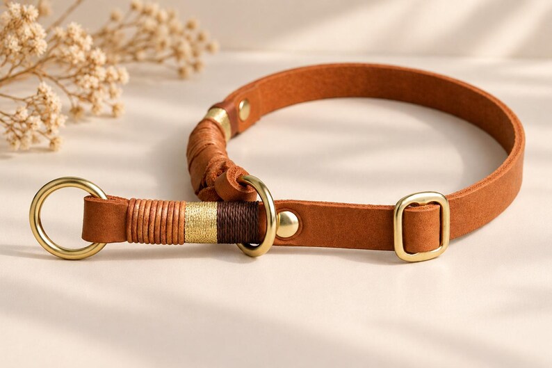 May include: A tan leather collar featuring brass hardware, including a circular ring and a rectangular buckle. The collar is accented with decorative thread wrapping in shades of tan, gold, and dark brown, secured with metal rivets.