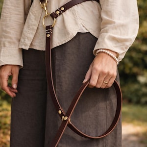 Op de afbeelding: Een handsfree hondenriem van donkerbruin leer met messing hardware, inclusief een ronde ringverbinding en verstelbare klinknagels. De riem is ontworpen om over het lichaam of rond de taille te worden gedragen met een lange band.