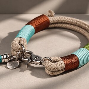 May include: A beige rope dog collar featuring sections wrapped in brown, turquoise, and green thread. It includes a silver metal lobster clasp, a matching turquoise tassel, and a round silver tag engraved with the text LIEBLINGS HUND.