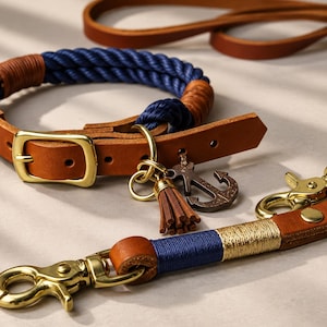 Op de afbeelding: Een halsband en riemset voor huisdieren met donkerblauw touw en bruine lederen accenten. De halsband heeft een messing gesp, een kleine lederen kwast en een ankerbedel. De riem is voorzien van messing musketonhaken en gouddraad.