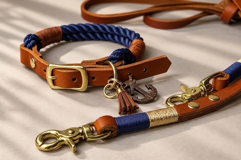 Op de afbeelding: Een halsband en riemset voor huisdieren met donkerblauw touw en bruine lederen accenten. De halsband heeft een messing gesp, een kleine lederen kwast en een ankerbedel. De riem is voorzien van messing musketonhaken en gouddraad.
