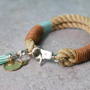 Op de afbeelding: Een beige touwarmband met secties omwikkeld met bruin leer en lichtblauw garen. De armband is voorzien van een zilverkleurige metalen karabijnsluiting, een kleine lichtblauwe kwast en een ronde goudkleurige metalen bedel met tekst.