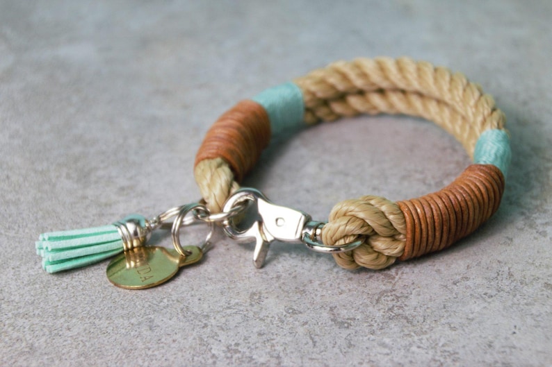 Op de afbeelding: Een beige touwarmband met secties omwikkeld met bruin leer en lichtblauw garen. De armband is voorzien van een zilverkleurige metalen karabijnsluiting, een kleine lichtblauwe kwast en een ronde goudkleurige metalen bedel met tekst.