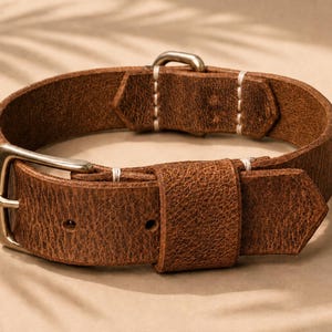 May include: A brown leather strap with a textured surface, featuring a metallic buckle and a matching metallic keeper. The strap is secured with white stitching at the connection points and includes two pre-punched holes for adjustment.