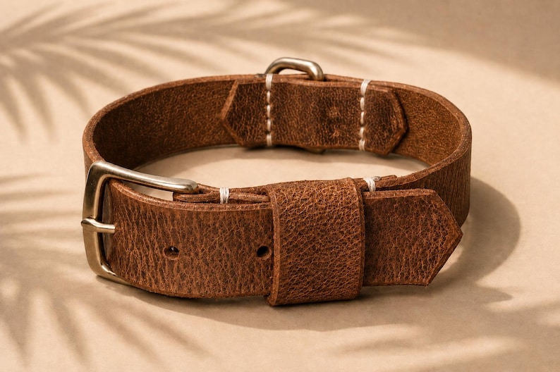 May include: A brown leather strap with a textured surface, featuring a metallic buckle and a matching metallic keeper. The strap is secured with white stitching at the connection points and includes two pre-punched holes for adjustment.