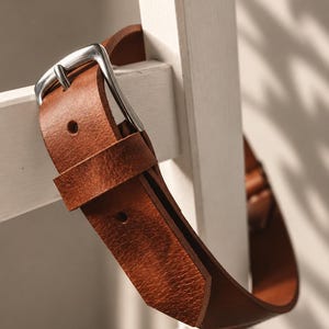 May include: A tan leather belt featuring a textured surface and a silver-toned metal buckle. The belt is constructed with a single leather loop and pre-punched holes for adjustment, showcasing a natural grain finish and visible stitching details.