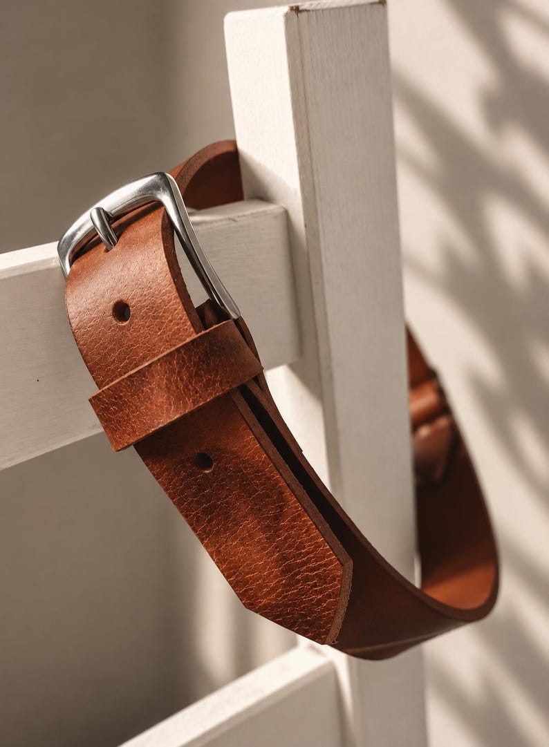 May include: A tan leather belt featuring a textured surface and a silver-toned metal buckle. The belt is constructed with a single leather loop and pre-punched holes for adjustment, showcasing a natural grain finish and visible stitching details.