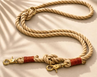 Op de afbeelding: Een beige touwlijn met twee goudkleurige metalen musketonhaken aan de uiteinden. Het touw is bij de bevestigingspunten vastgezet met decoratief koperkleurig garen en is voorzien van een vaste knoop voor een verstelbare lengte.