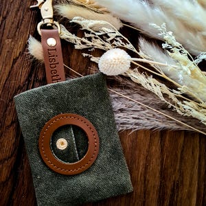 Könnte beinhalten: Ein grüner Hundekotbeutelhalter aus Canvas mit einem braunen Leder-Ring und einem braunen Lederband mit dem eingravierten Namen "Lisbeth". Der Halter ist an einem goldenen Metallclip befestigt.