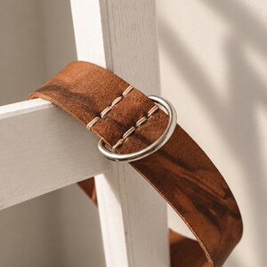 May include: A brown leather belt featuring a smooth, matte finish with visible dark grain patterns. The belt is secured with a silver-toned metal D-ring buckle and is accented by thick, light-colored stitching near the closure point.