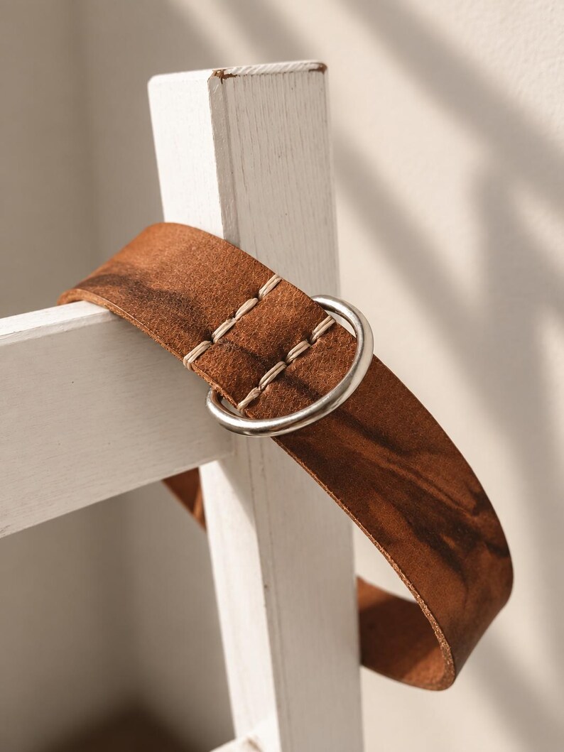 May include: A brown leather belt featuring a smooth, matte finish with visible dark grain patterns. The belt is secured with a silver-toned metal D-ring buckle and is accented by thick, light-colored stitching near the closure point.