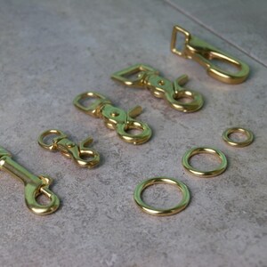 Op de afbeelding: Een verzameling goudkleurige metalen hardware-onderdelen, waaronder vier draaibare karabijnsluitingen van verschillende groottes en drie ronde ringen met vari&euml;rende diameters, allemaal met een gladde, gepolijste en reflecterende afwerking.