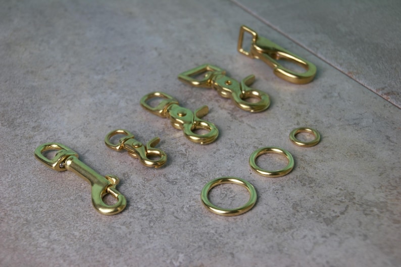 Op de afbeelding: Een verzameling goudkleurige metalen hardware-onderdelen, waaronder vier draaibare karabijnsluitingen van verschillende groottes en drie ronde ringen met vari&euml;rende diameters, allemaal met een gladde, gepolijste en reflecterende afwerking.