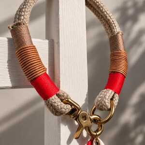 May include: A beige rope collar featuring decorative wrappings in brown, tan, and bright red thread. The collar includes a brass swivel snap hook and a brass ring, with a small red tassel and a white cowrie shell charm attached to the hardware.