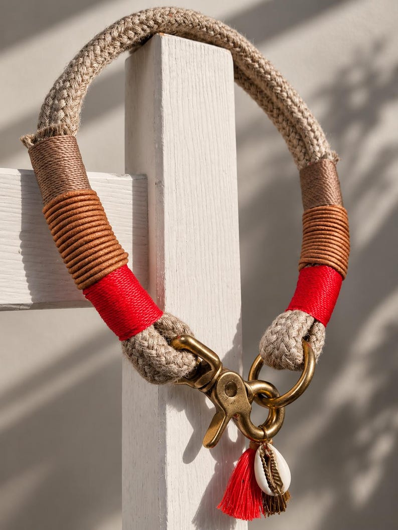 May include: A beige rope collar featuring decorative wrappings in brown, tan, and bright red thread. The collar includes a brass swivel snap hook and a brass ring, with a small red tassel and a white cowrie shell charm attached to the hardware.