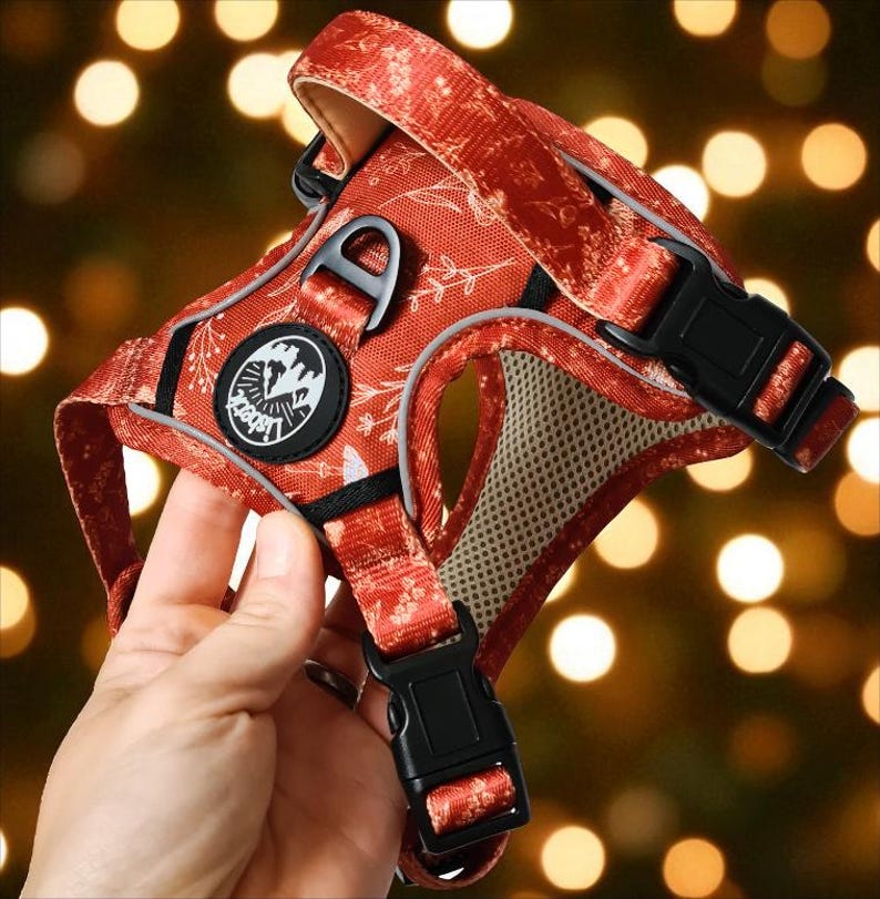 May include: A rust-orange pet harness featuring a white botanical floral pattern, black plastic side-release buckles, and a circular black patch with white mountain graphics and the text Lisbet. The interior is lined with beige mesh fabric.