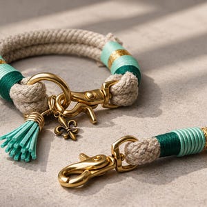 May include: A beige rope collar and matching leash attachment featuring gold-toned metal hardware, including a lobster clasp and a fleur-de-lis charm. The rope is accented with wrapped thread in shades of mint green, dark green, and metallic gold.
