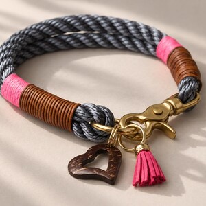 Op de afbeelding: Een halsband van gedraaid grijs touw met secties omwikkeld met felroze draad en bruin koord. Het bevat een messingkleurige metalen karabijnsluiting, een donkerbruine houten hartvormige bedel en een klein roze kwastje aan een ring.