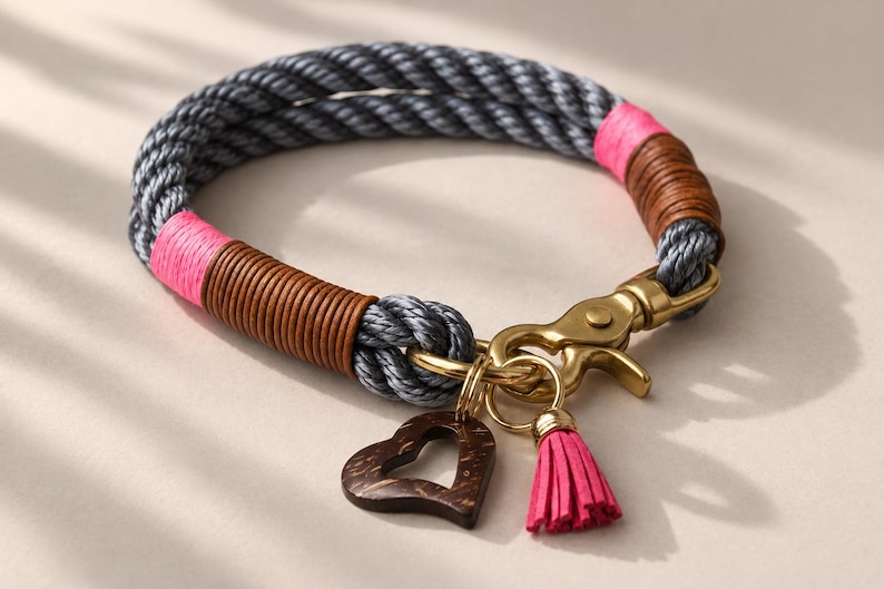 Op de afbeelding: Een halsband van gedraaid grijs touw met secties omwikkeld met felroze draad en bruin koord. Het bevat een messingkleurige metalen karabijnsluiting, een donkerbruine houten hartvormige bedel en een klein roze kwastje aan een ring.