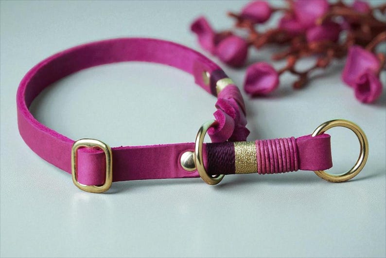 May include: A bright pink leather collar featuring gold-toned metal hardware, including a rectangular buckle and two circular rings. The strap is accented with decorative thread wrapping in shades of dark purple, gold, and pink near the ring.