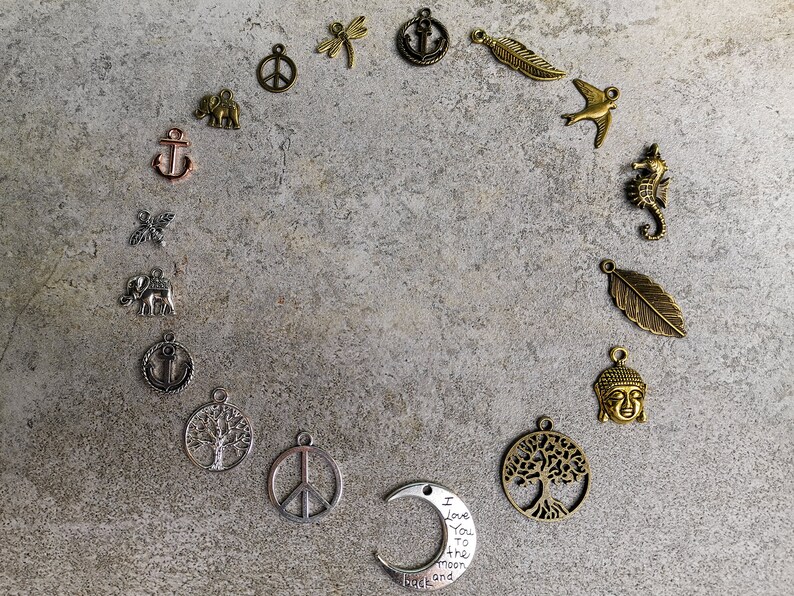Pode incluir: Uma cole&ccedil;&atilde;o de berloques de metal dispostos em c&iacute;rculo, incluindo formas de &acirc;ncoras, elefantes, s&iacute;mbolos da paz, lib&eacute;lulas, penas, p&aacute;ssaros, cavalos-marinhos, &aacute;rvores, cabe&ccedil;as de Buda e um berloque de meia-lua com o texto I Love You to the moon and back.