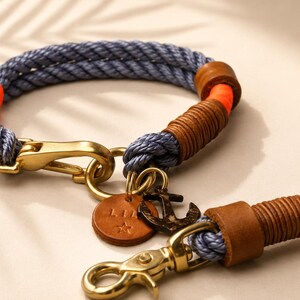 Op de afbeelding: Een blauwe touwlijn en halsbandset met feloranje accenten, bruine lederen wikkelingen en goudkleurige metalen hardware. De set bevat een ronde leren tag met de tekst LIA en een ster, samen met een donker metalen ankerbedel.