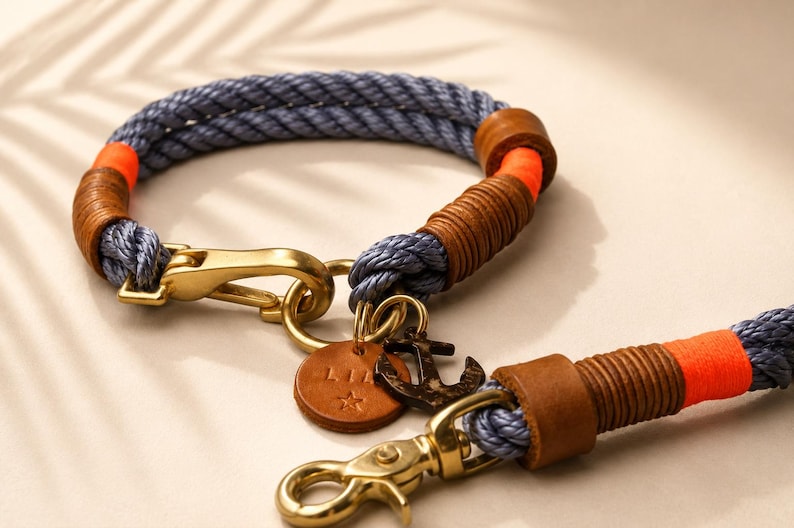 Op de afbeelding: Een blauwe touwlijn en halsbandset met feloranje accenten, bruine lederen wikkelingen en goudkleurige metalen hardware. De set bevat een ronde leren tag met de tekst LIA en een ster, samen met een donker metalen ankerbedel.