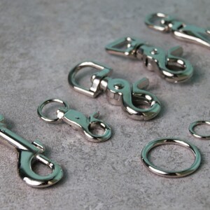 Op de afbeelding: Een verzameling zilverkleurige metalen draaibare karabijnsluitingen in verschillende maten, voorzien van veerbelaste sluitingen en roterende basissen, vergezeld door twee ronde metalen ringen van verschillende diameters.