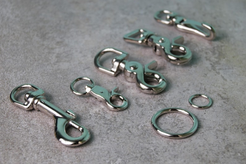 Op de afbeelding: Een verzameling zilverkleurige metalen draaibare karabijnsluitingen in verschillende maten, voorzien van veerbelaste sluitingen en roterende basissen, vergezeld door twee ronde metalen ringen van verschillende diameters.