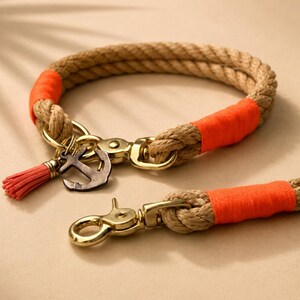 Op de afbeelding: Een halsband en riem van beige touw met feloranje draadomwikkelingen, goudkleurige metalen hardware en een kleine metalen ankerbedel gecombineerd met een bijpassend oranje kwastje aan de ring van de halsband.