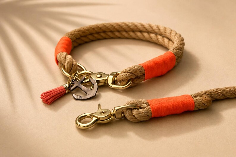Op de afbeelding: Een halsband en riem van beige touw met feloranje draadomwikkelingen, goudkleurige metalen hardware en een kleine metalen ankerbedel gecombineerd met een bijpassend oranje kwastje aan de ring van de halsband.