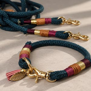 May include: A dark teal rope lead and matching collar set featuring gold-tone metal hardware, decorative maroon and tan thread wrappings, and a small gold leaf charm with a pink tassel attached to the collar's metal clasp mechanism.