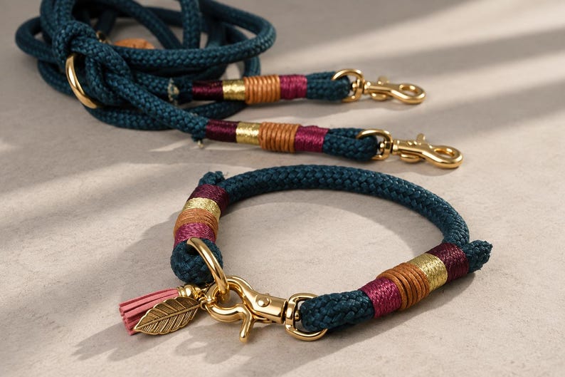 May include: A dark teal rope lead and matching collar set featuring gold-tone metal hardware, decorative maroon and tan thread wrappings, and a small gold leaf charm with a pink tassel attached to the collar's metal clasp mechanism.