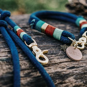May include: A dark blue rope dog collar and matching lead set featuring brass hardware, decorative teal, red, and gold thread wrappings, a small metal star charm, and a round tan leather tag with the embossed name LULU attached to the collar.