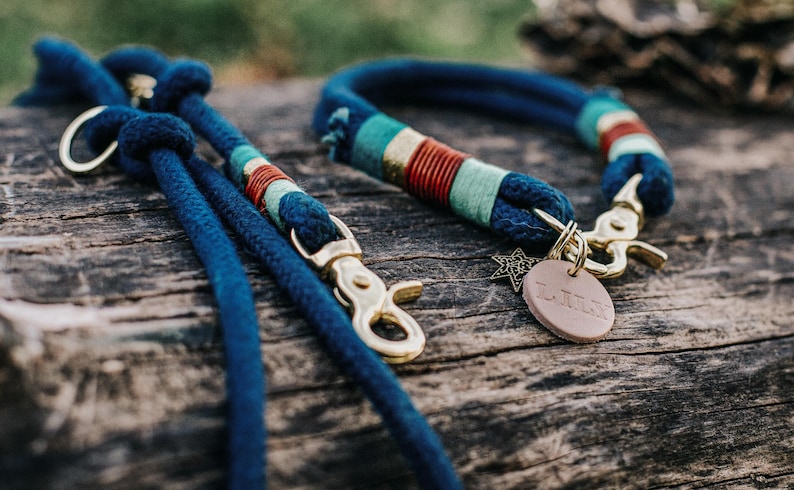 May include: A dark blue rope dog collar and matching lead set featuring brass hardware, decorative teal, red, and gold thread wrappings, a small metal star charm, and a round tan leather tag with the embossed name LULU attached to the collar.