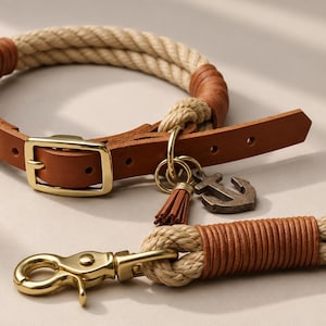 Op de afbeelding: Een halsband en riemset voor huisdieren gemaakt van beige touw met lichtbruine lederen accenten. De halsband is voorzien van een goudkleurige metalen gesp en een metalen ankerbedel met een bijpassend leren kwastje.