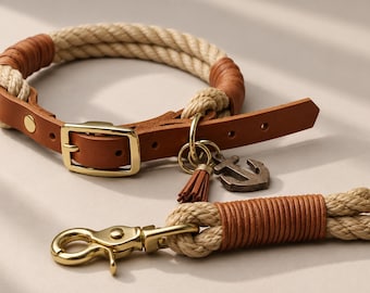 Op de afbeelding: Een halsband en riemset voor huisdieren gemaakt van beige touw met lichtbruine lederen accenten. De halsband is voorzien van een goudkleurige metalen gesp en een metalen ankerbedel met een bijpassend leren kwastje.