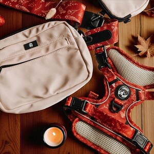 May include: A beige rectangular crossbody bag with a black zipper and a small black logo patch, paired with a red pet harness featuring a white floral pattern, black buckles, and a circular logo patch with a mountain graphic and the text "Lishinu".
