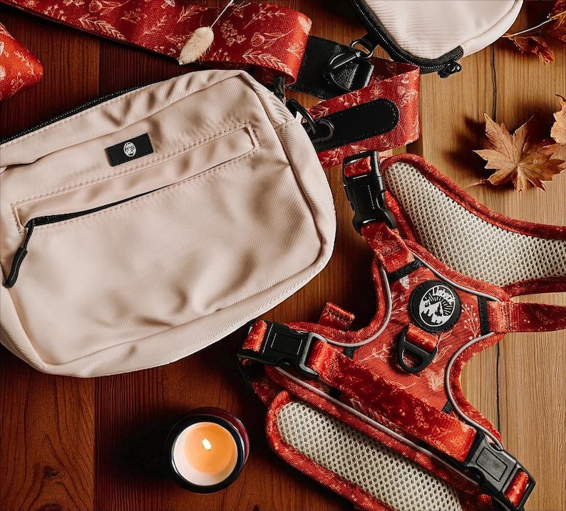 May include: A beige rectangular crossbody bag with a black zipper and a small black logo patch, paired with a red pet harness featuring a white floral pattern, black buckles, and a circular logo patch with a mountain graphic and the text "Lishinu".