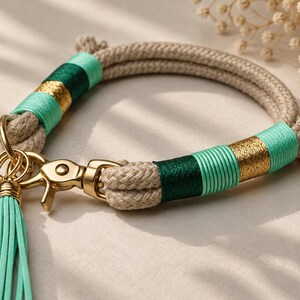 May include: A beige rope bracelet featuring sections wrapped in mint green, dark green, and metallic gold thread. The bracelet includes a gold-toned metal lobster clasp, a matching peace sign charm, and a long mint green faux leather tassel.