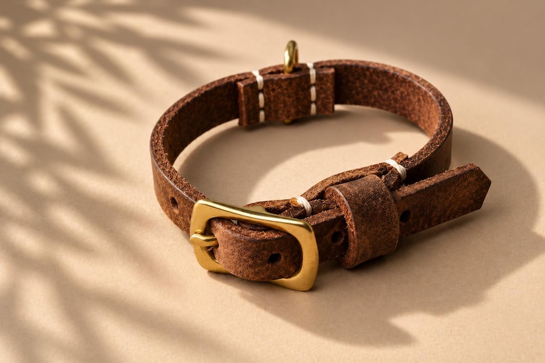 May include: A dark brown leather collar featuring a polished gold-tone metal buckle and a matching D-ring. The strap is constructed with visible white stitching at the connection points and includes a sliding leather keeper loop for adjustment.